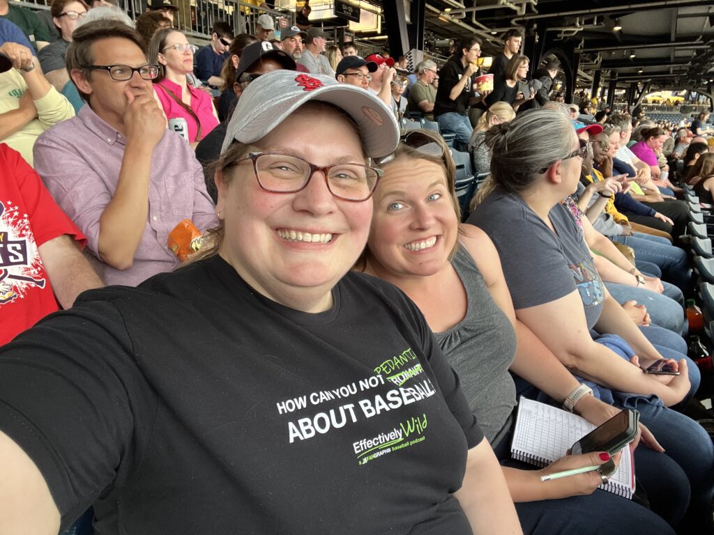 a white woman grins at the camera wearing a shirt that reads, "how can you not be pedantic about baseball?"; she is surrounded by a crowd of people watching a baseball game